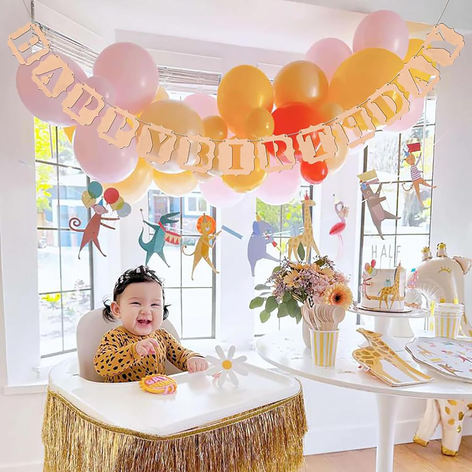 Child in a high chair with 'Happy Birthday' banner and party decorations in a room with large windows.