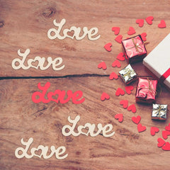 Wooden surface with 'love' word cutouts, small gift boxes, and red heart-shaped confetti.
