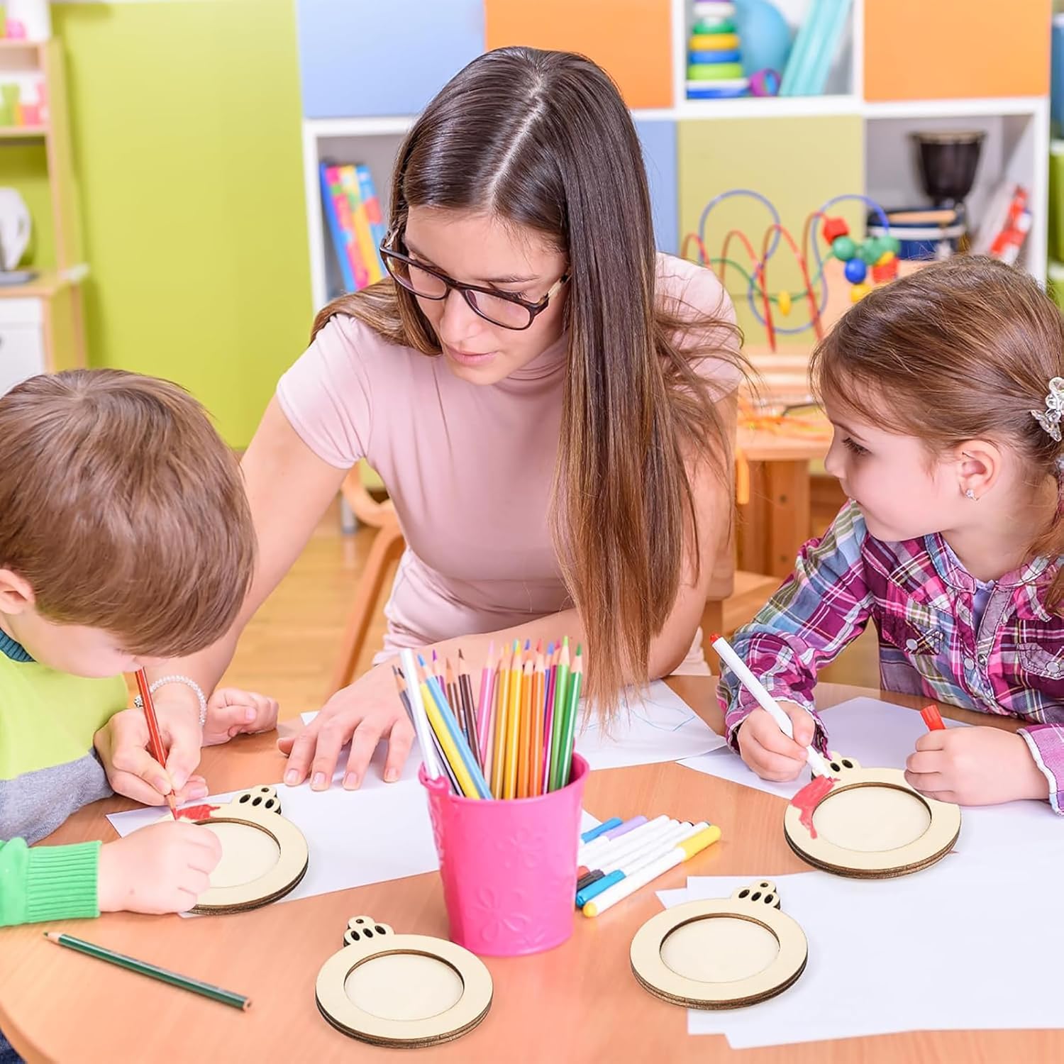 Teacher assisting two children with art projects in a classroom setting.