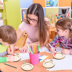 Teacher assisting two children with art projects in a classroom setting.