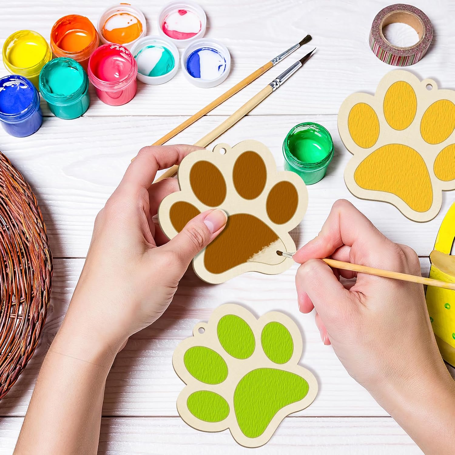Hands painting wooden paw prints with colorful paint and brushes on a white surface.