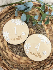 Two wooden milestone markers with floral designs and numbers on a woven mat.