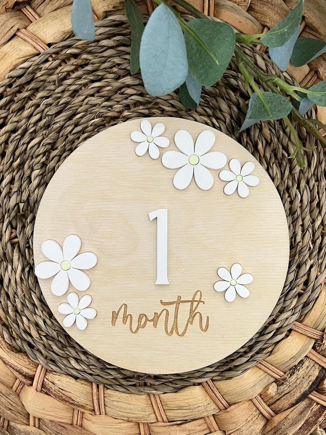 Round wooden plaque with '1 month' and white flowers on a woven surface with greenery.