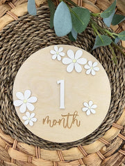 Round wooden plaque with '1 month' and white flowers on a woven surface with greenery.