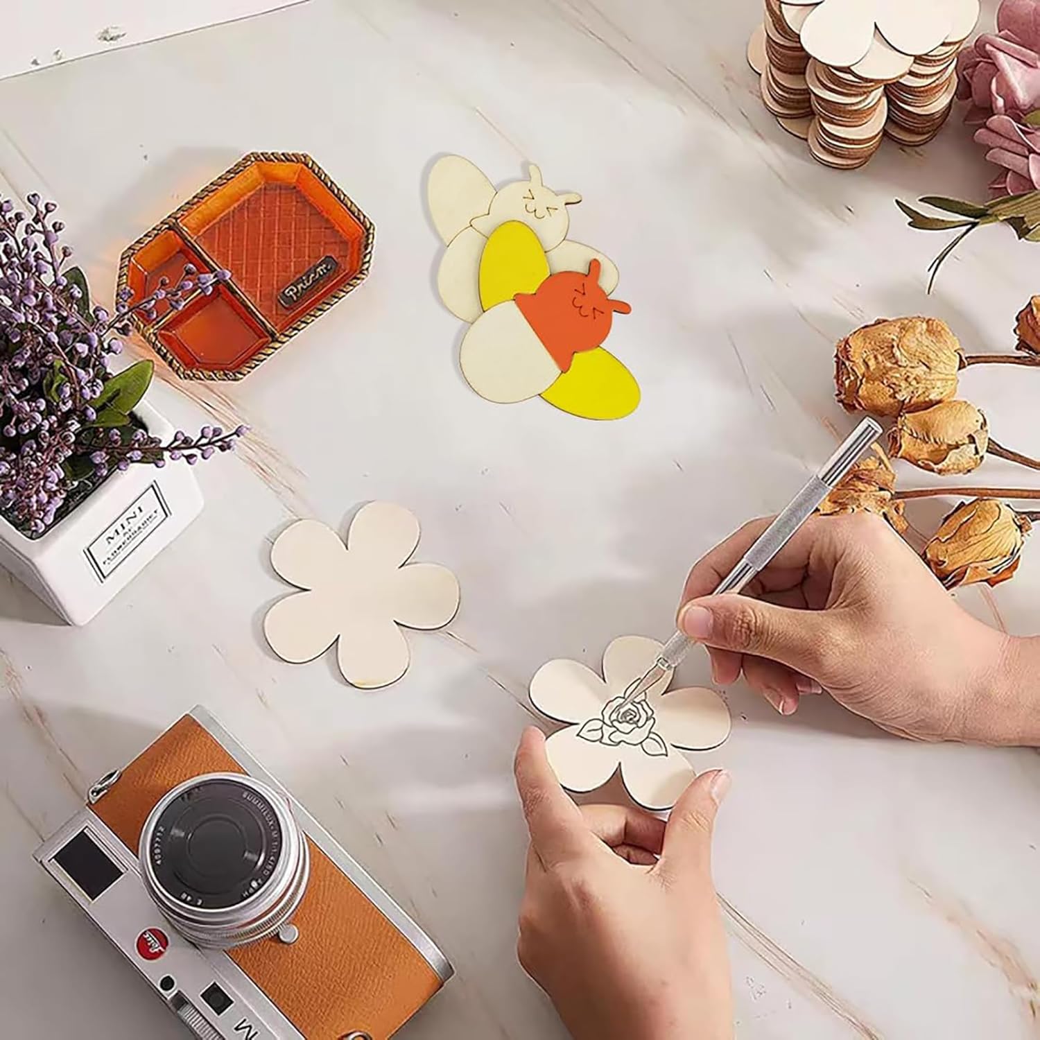 Person decorating a white flower-shaped coaster with a pen on a table with a camera and decorative items.
