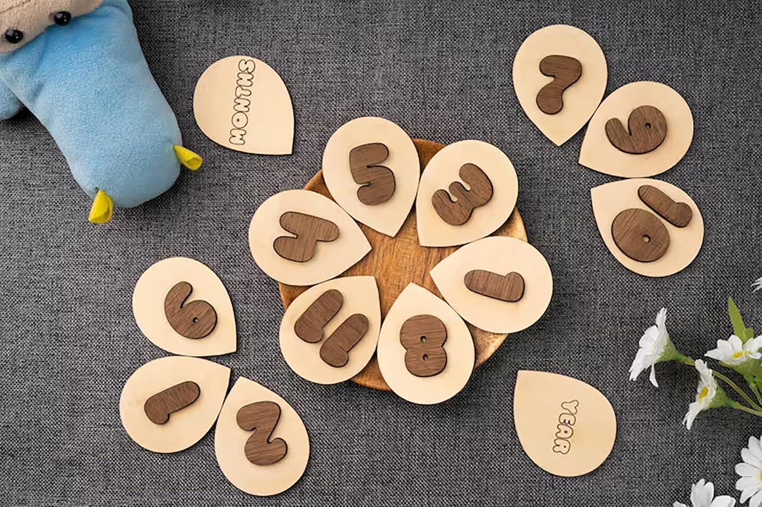 Wooden numbers on a circular wooden board with a plush toy and flowers in the background.