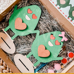 Green paper cutouts of a face with heart-shaped eyes and mouth on straw, with wooden tools and pink and red items in the background.