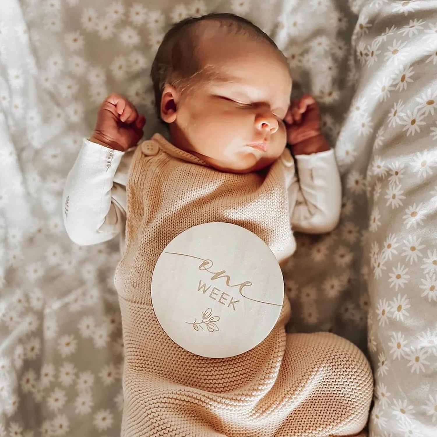 Newborn baby wearing a beige outfit with a 'One Week' milestone card, lying on a floral blanket.