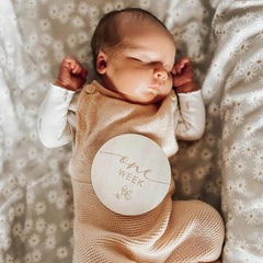 Newborn baby wearing a beige outfit with a 'One Week' milestone card, lying on a floral blanket.