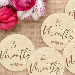 Wooden milestone discs with month counts on a textured surface with flowers.