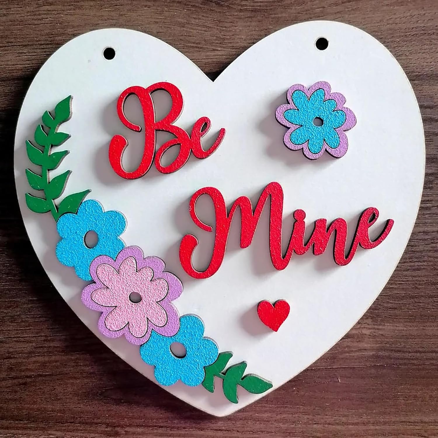 Heart-shaped decorative sign with 'Be Mine' text and colorful flowers on a wooden background