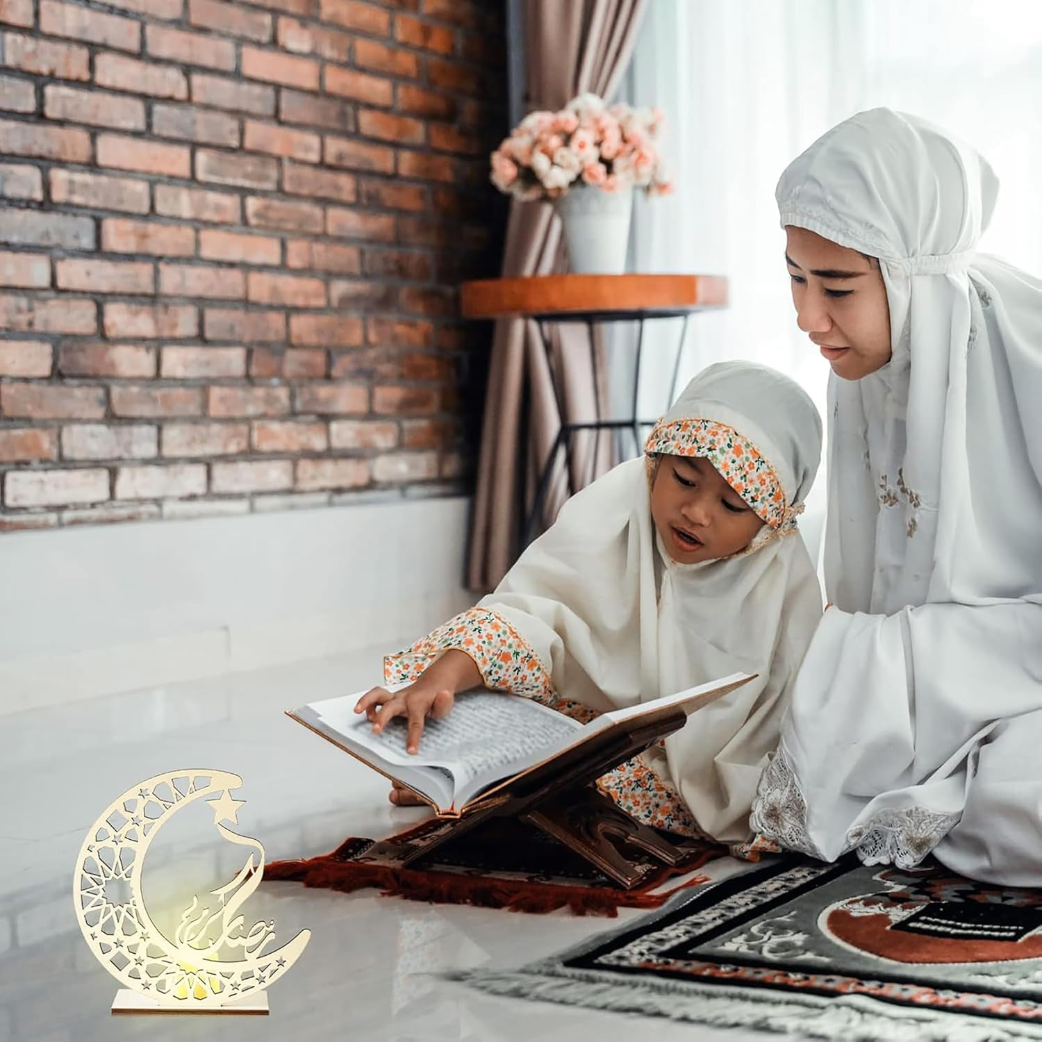 Two women in hijabs reading a book together with a decorative moon lamp on a table.