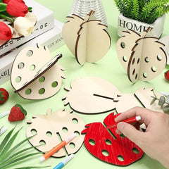 Wooden strawberry shapes with a hand using a red marker on a green surface with flowers and books.