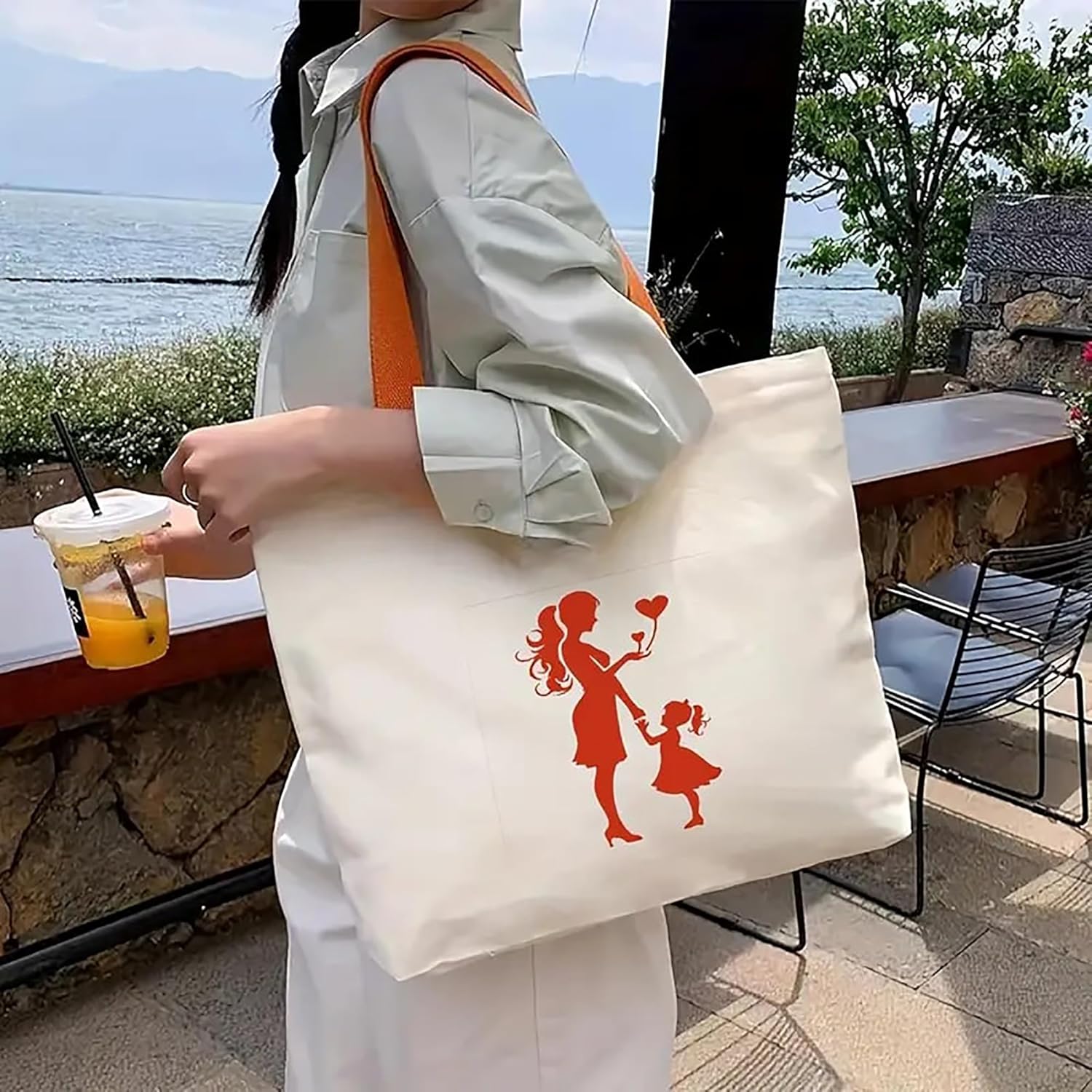Person holding a tote bag with a design of a woman and child, outdoors by a body of water.