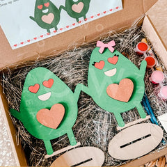 Green paper craft figures with heart shapes on a bed of straw, with a box in the background.