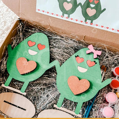 Green paper cutouts of cartoon characters with hearts, one wearing a bow, in a box with craft supplies.