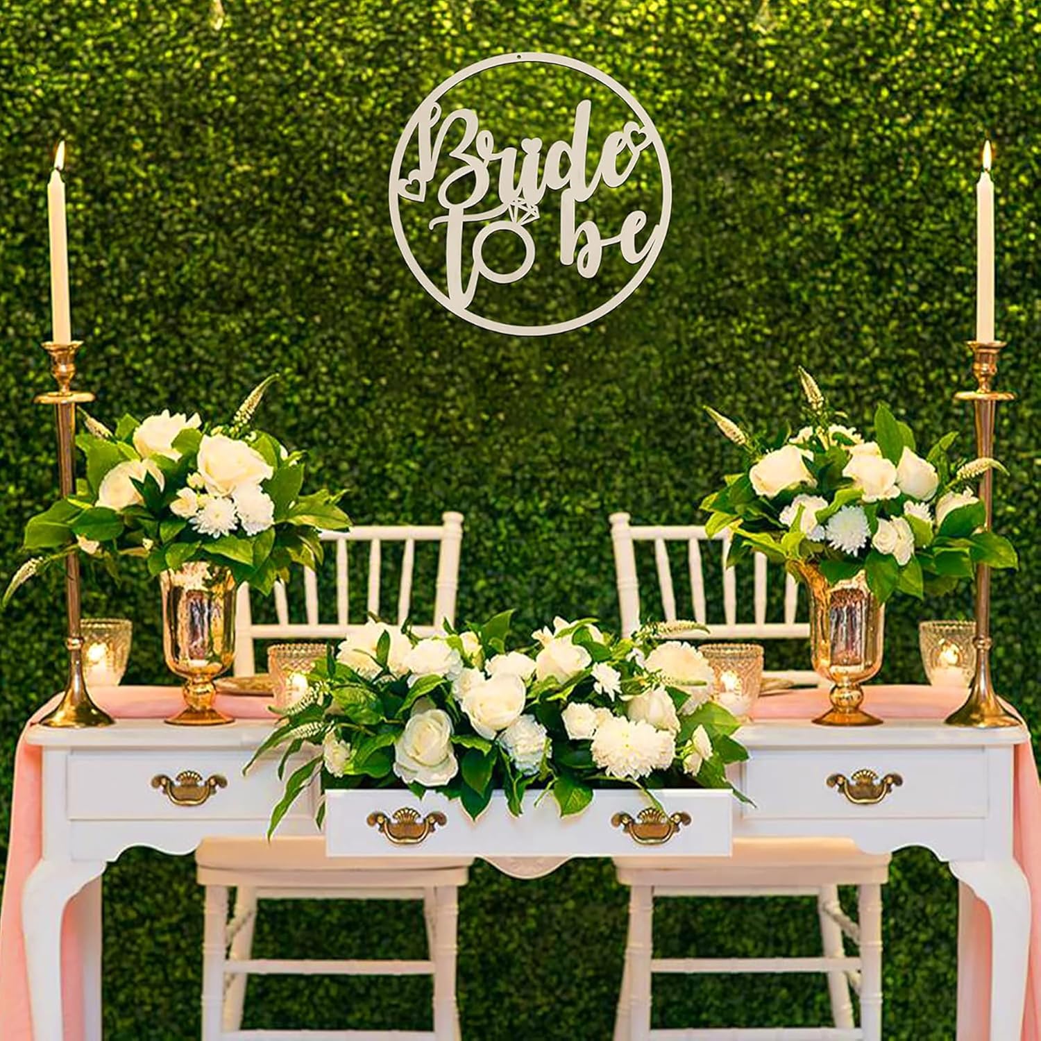 Decorated table with floral arrangements, candles, and 'Bride to be' sign against a green hedge background.