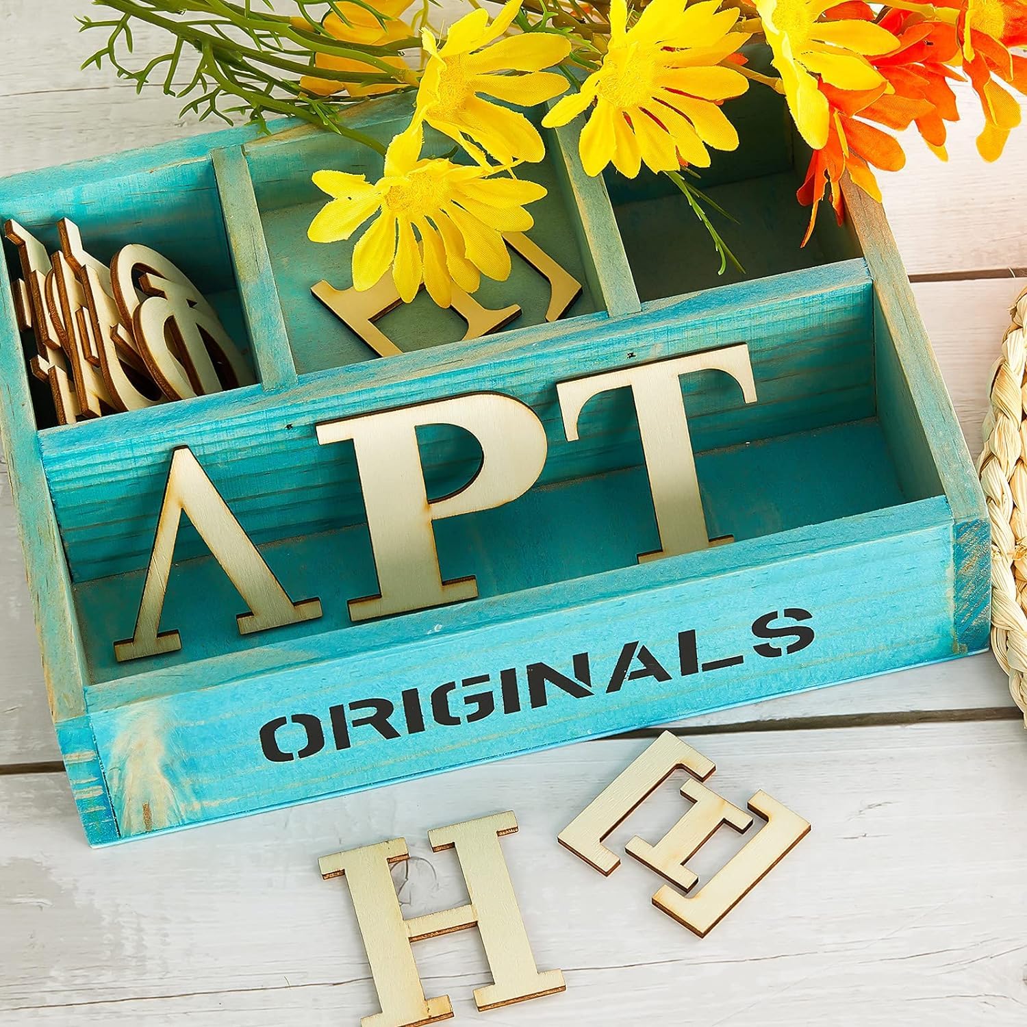 Teal wooden box with gold letters spelling 'APT' and 'ORIGINALS', surrounded by yellow flowers on a light wooden surface.