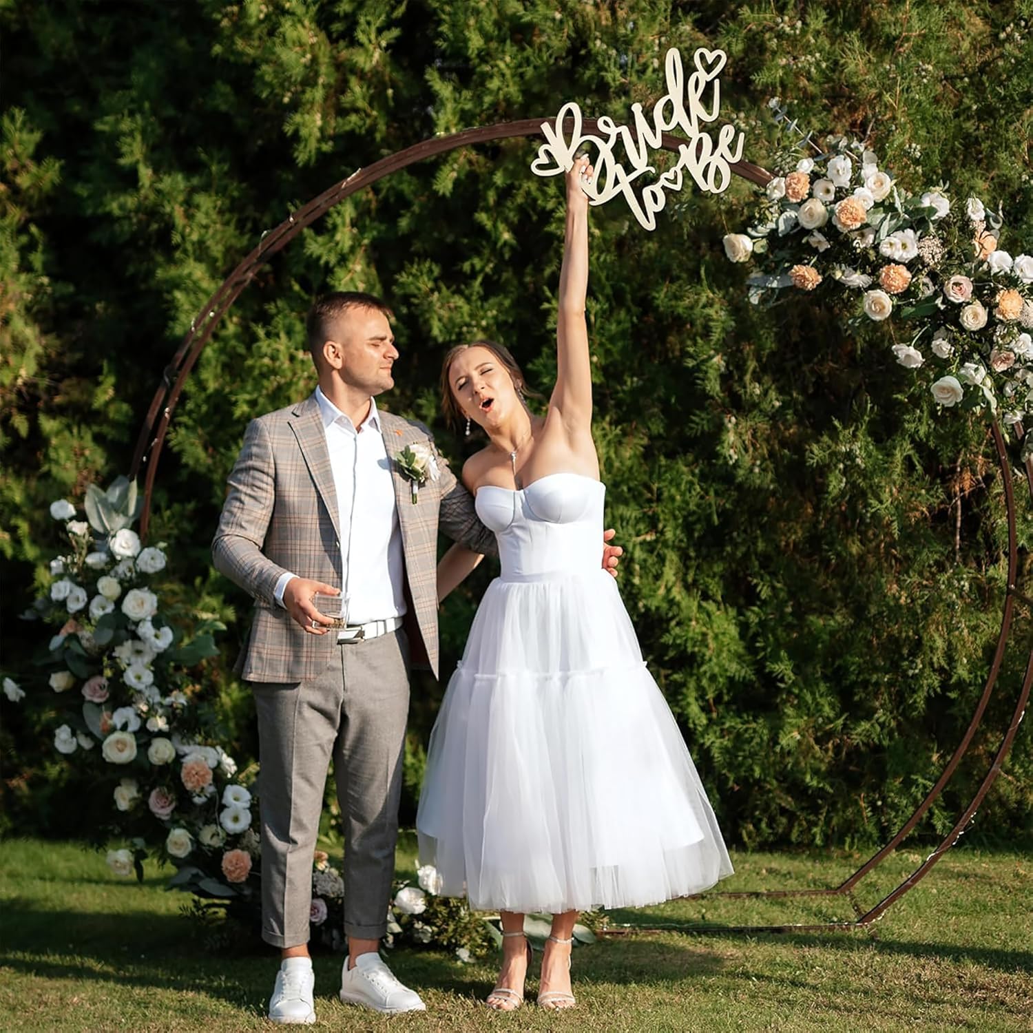 Couple standing under a decorative arch with floral arrangements on a sunny day.