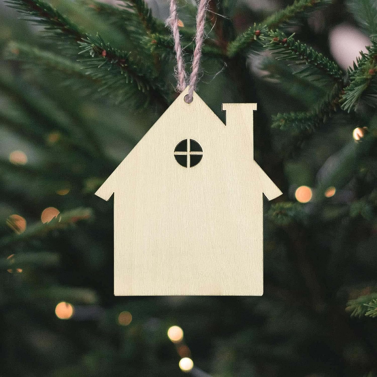 Wooden house-shaped ornament hanging on a Christmas tree with blurred lights in the background.
