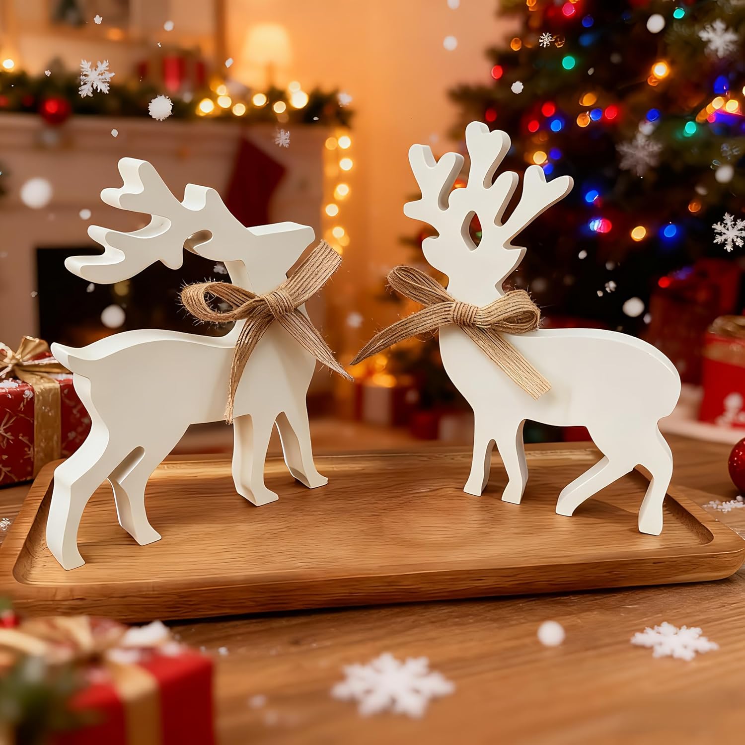 Two white deer figurines with brown bows on a wooden tray in a festive setting with Christmas tree and presents.