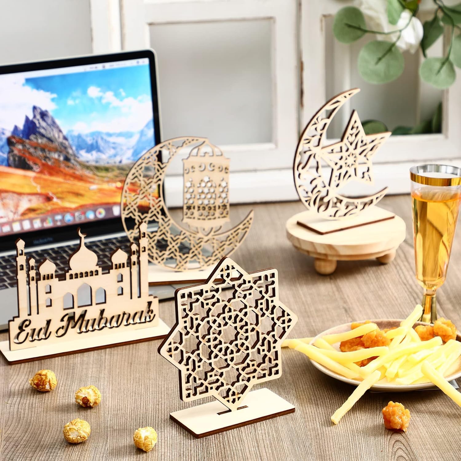 Decorative wooden 'Eid Mubarak' items on a table with food and a laptop in the background.