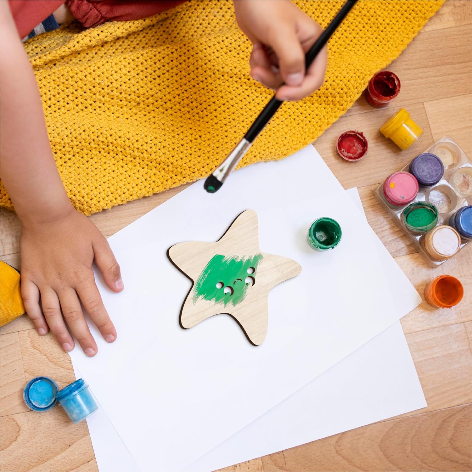 Child painting a star-shaped wooden object with colorful paint on a white sheet of paper.