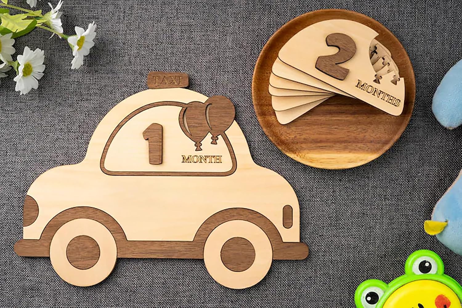 Wooden car-shaped milestone with number '1' and wooden plate with number '2', on a textured surface with flowers and a frog toy.