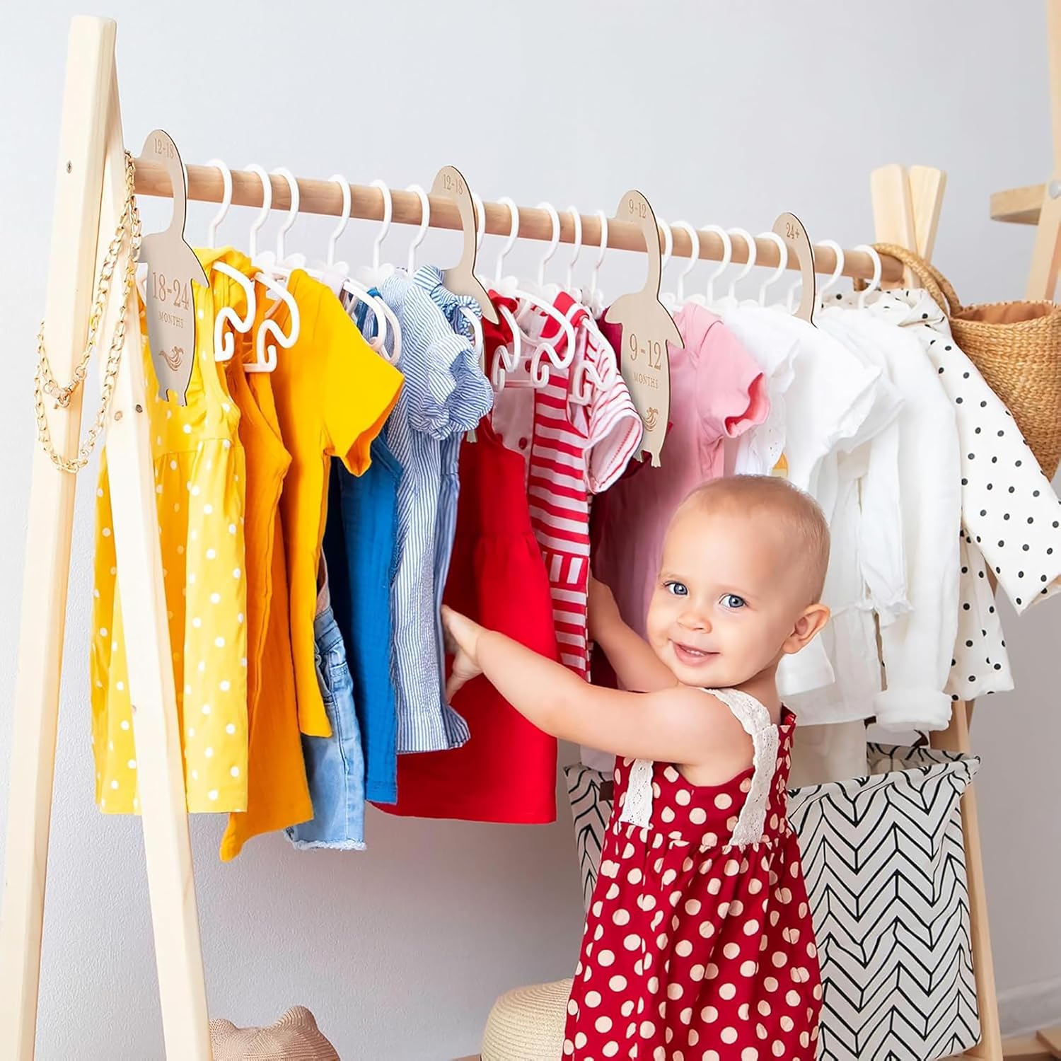 Baby in a red polka dot dress standing next to a rack of colorful clothes.