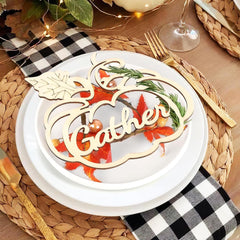 Decorative place setting with 'Gather' sign on a white plate, surrounded by checkered napkin, leaves, and wine glass.
