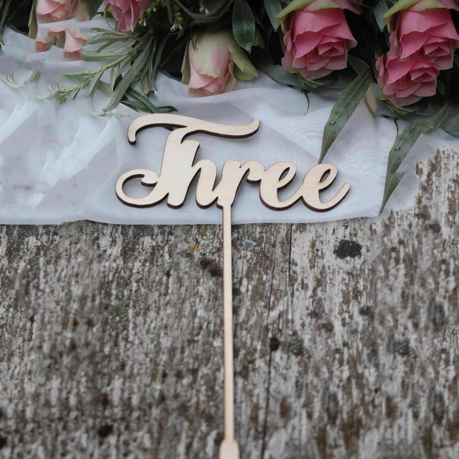 Wooden 'Three' cake topper with pink roses and greenery on a rustic wooden surface