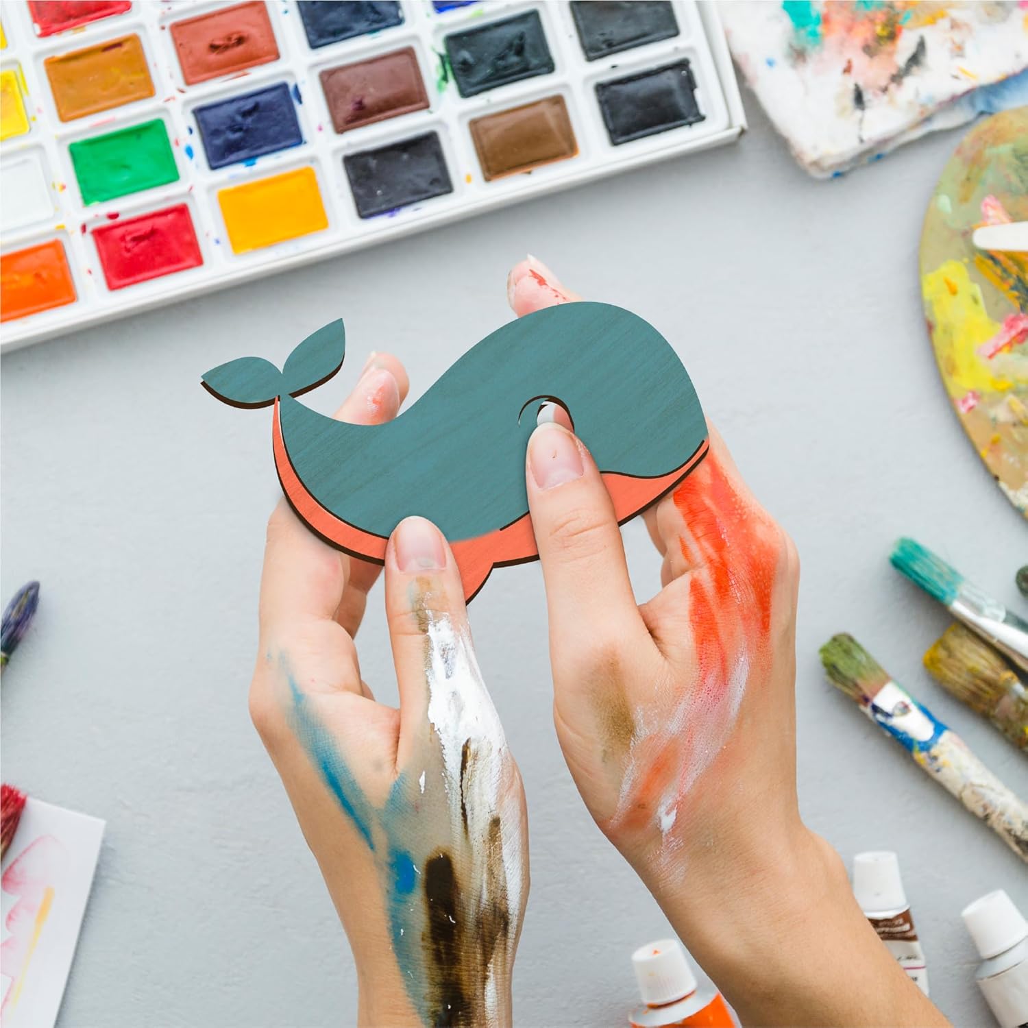 Hands holding a whale-shaped object with watercolor paints and brushes on a gray surface