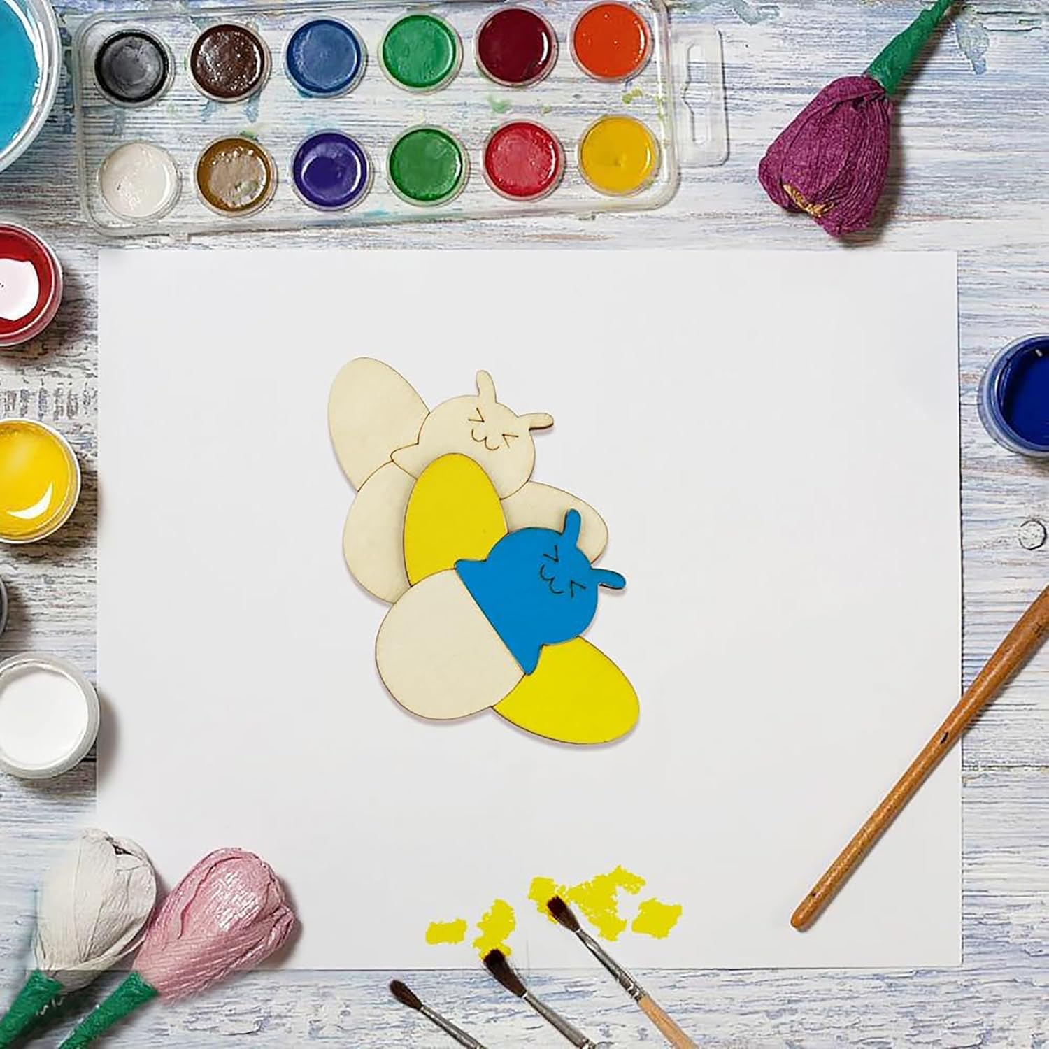 Colorful bee-shaped cookie on a white sheet of paper with paint supplies around.