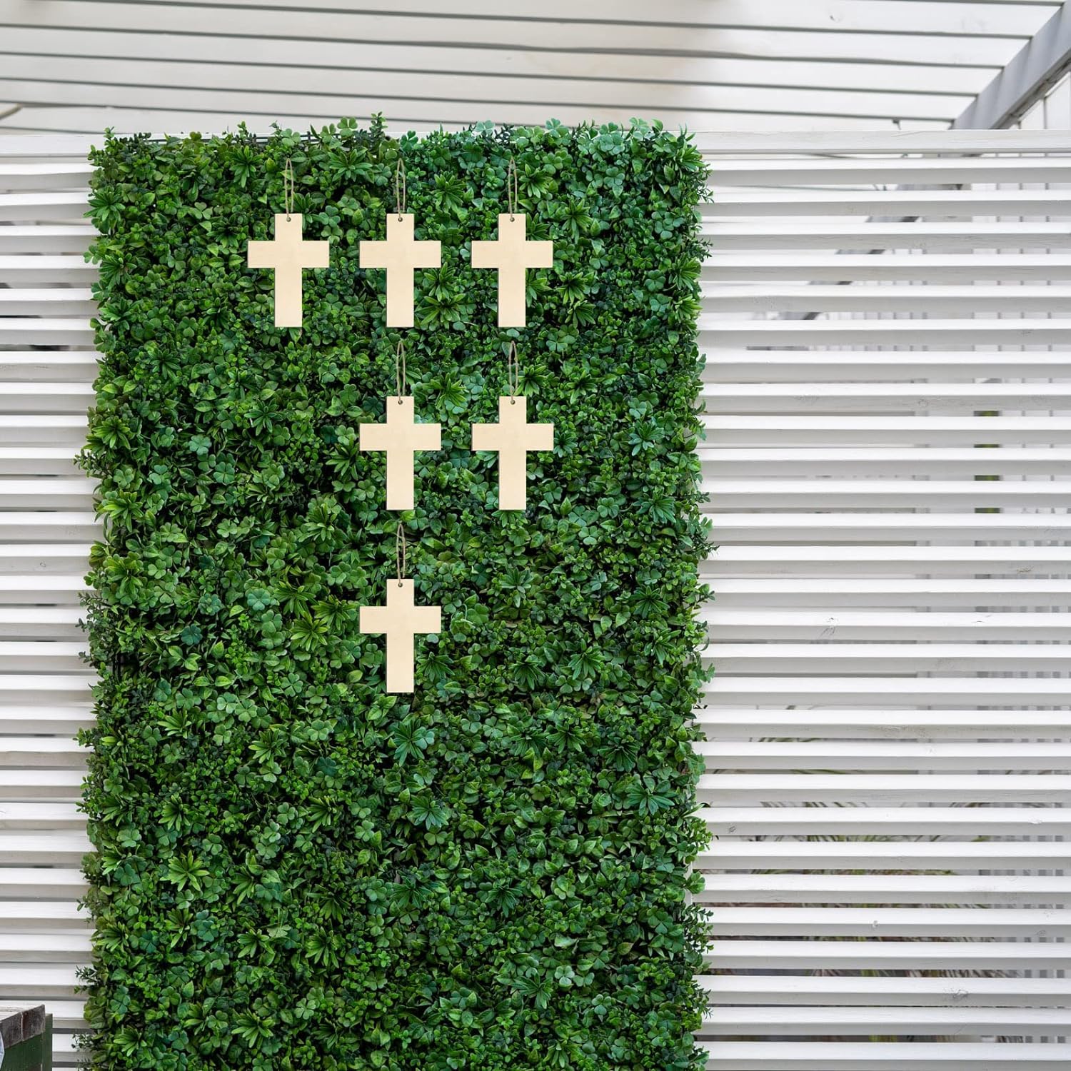 Green artificial hedge wall with wooden crosses against a white slatted background