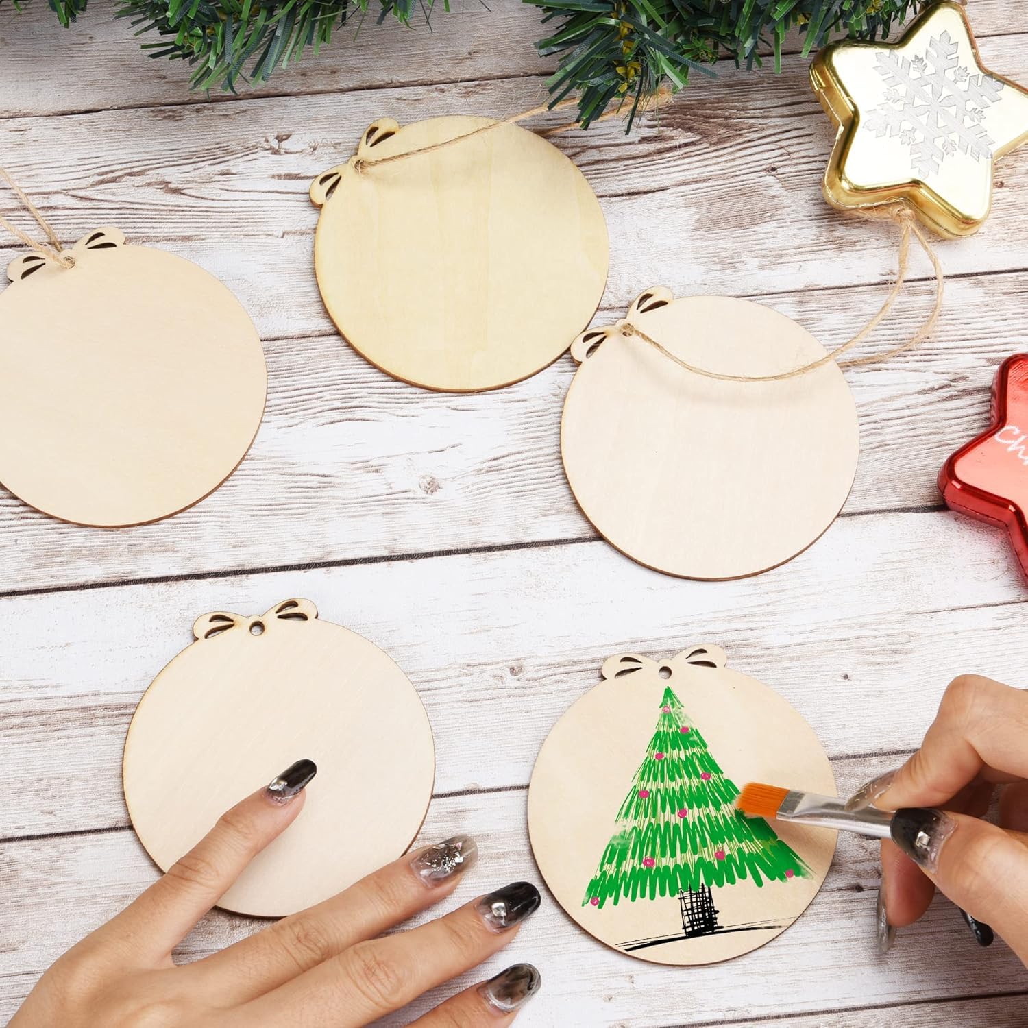 Person painting a Christmas tree on a wooden ornament with other ornaments and decorative items in the background.