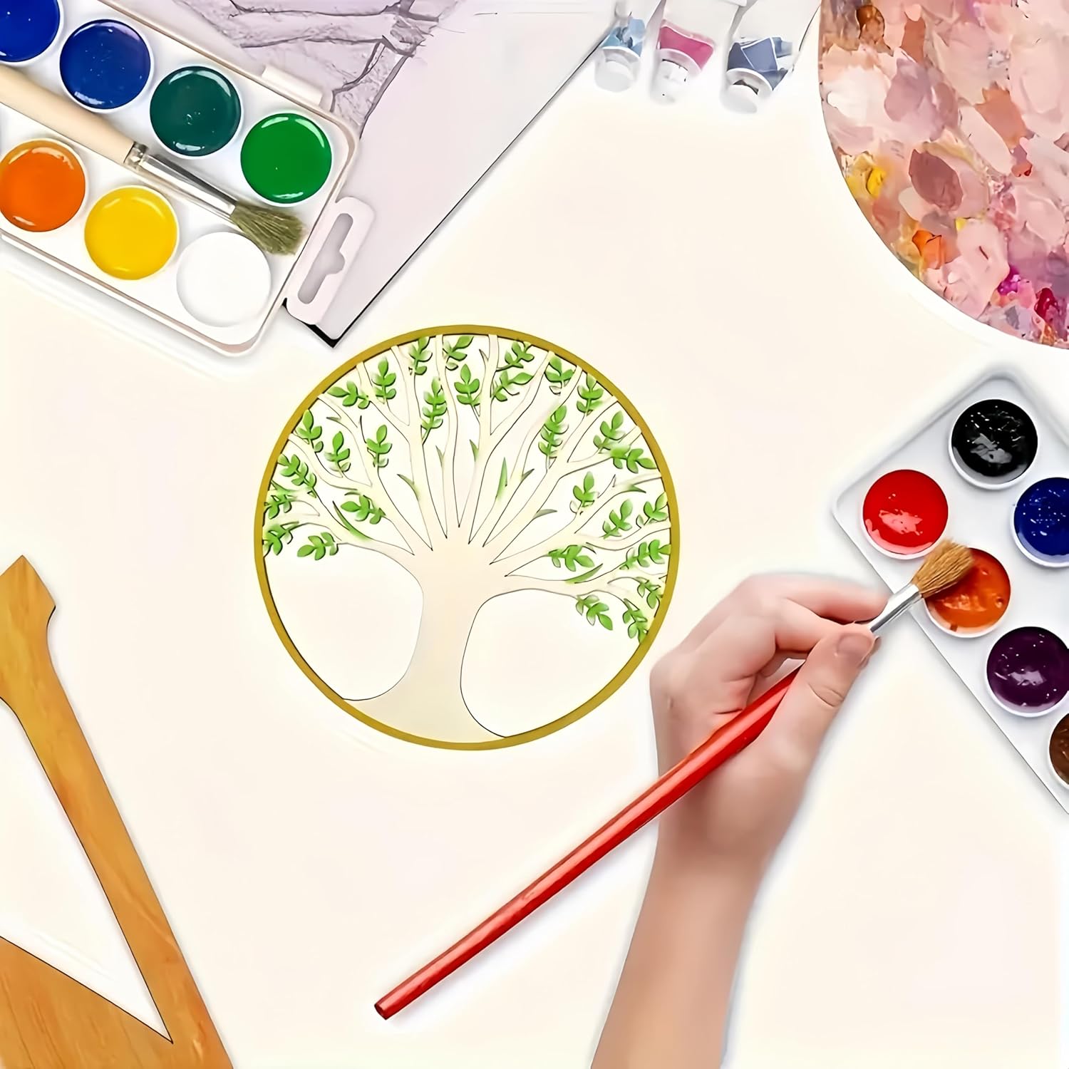 Artistic setup with watercolor paints, a palette, and a hand holding a brush near a painted tree design.