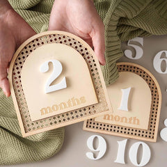 Wooden milestone cards with numbers held by a hand, against a green textured fabric background.