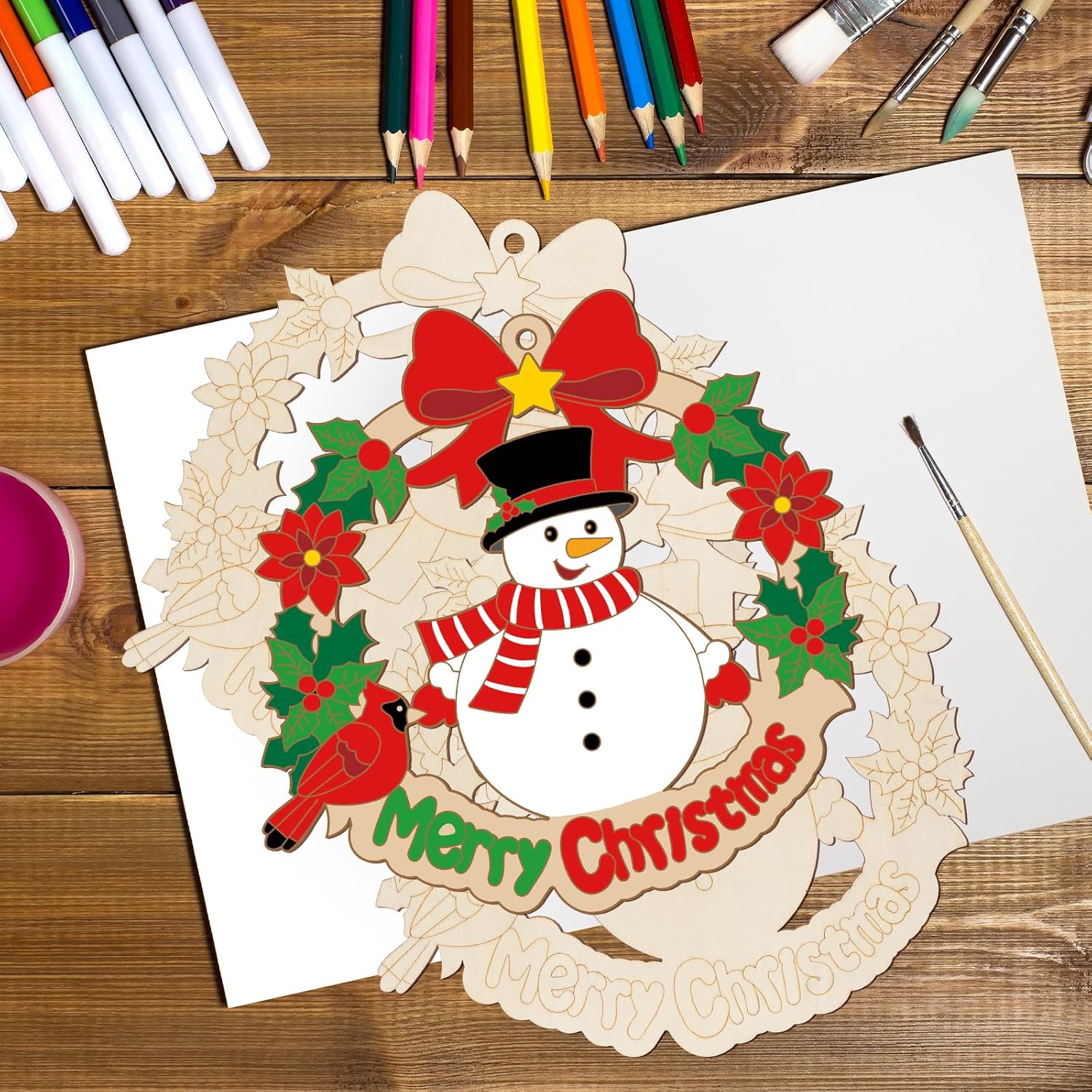 Christmas-themed wooden cutout with a snowman, wreath, and 'Merry Christmas' text on a wooden surface with coloring supplies.