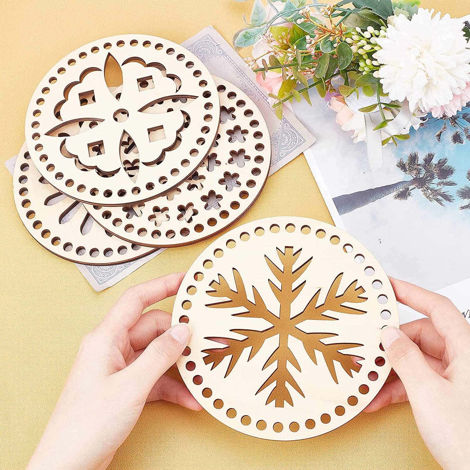 Wooden coasters with snowflake and floral designs held by hands on a yellow surface with flowers in the background.