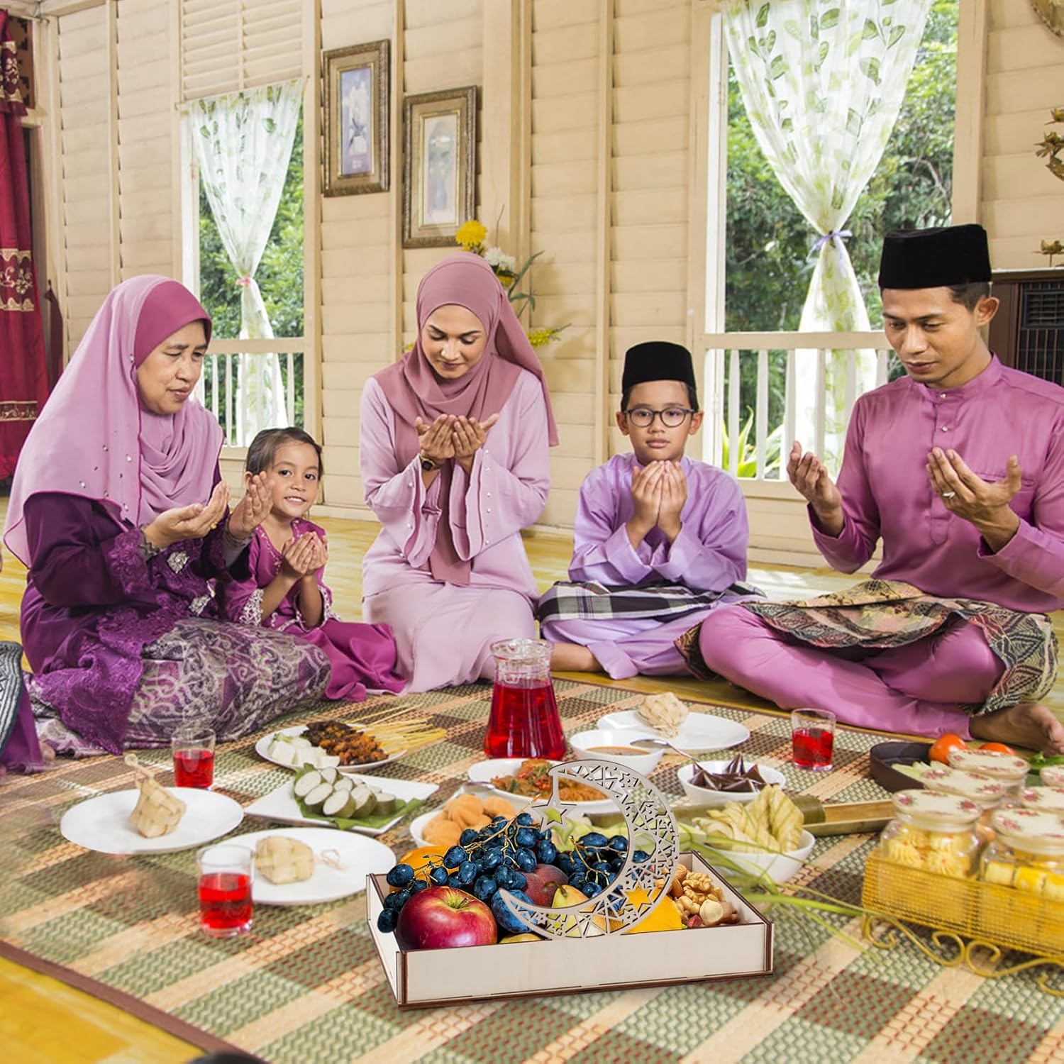 Family praying together during a meal with fruits and drinks on a patterned rug.