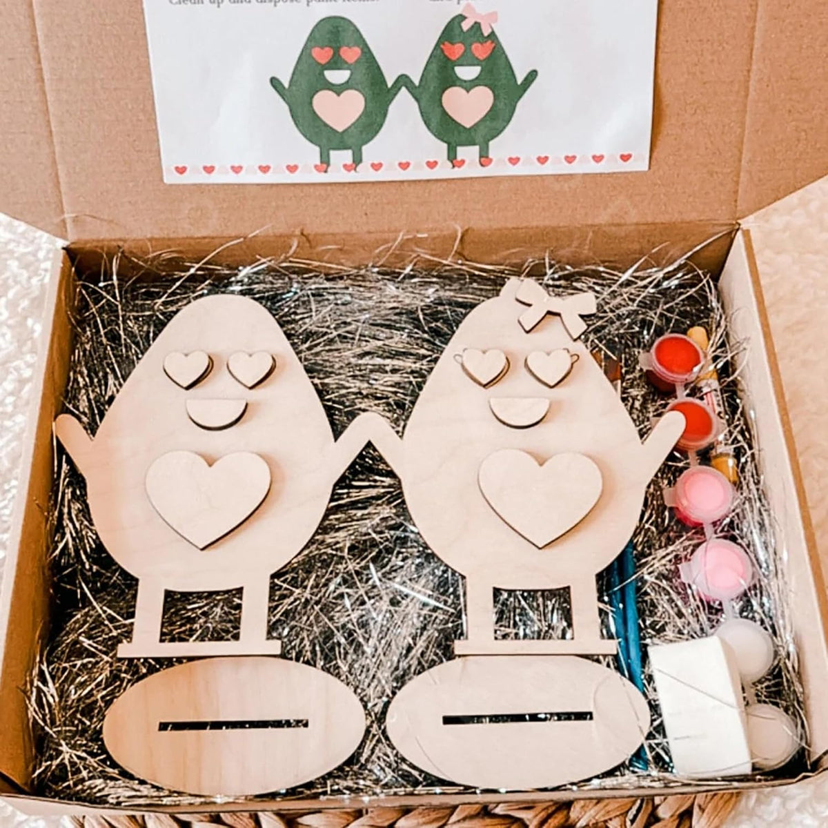 Wooden egg-shaped characters with heart designs in a box with straw padding.