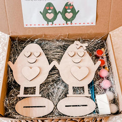 Wooden egg-shaped characters with heart designs in a box with straw padding.