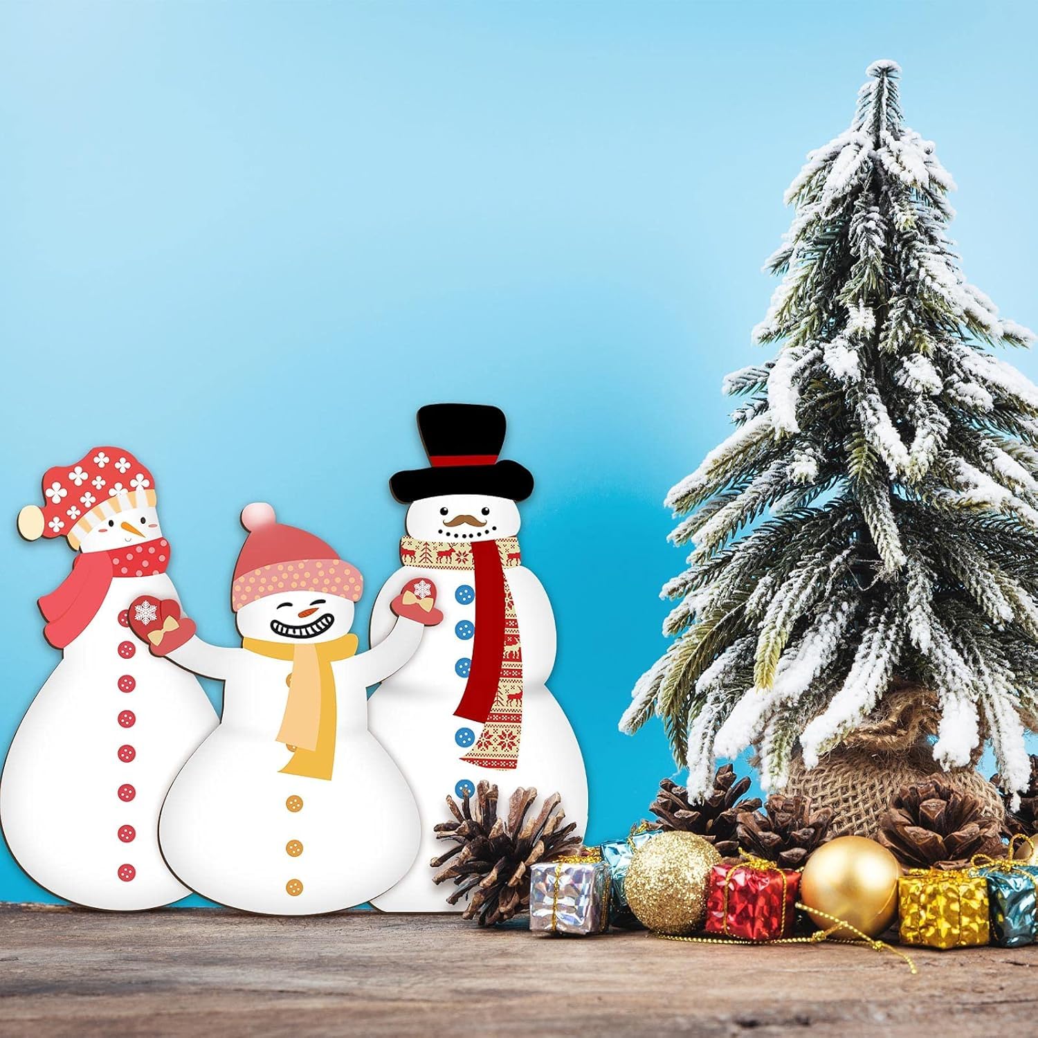 Three snowmen with colorful hats and scarves next to a small Christmas tree with decorations on a wooden surface.