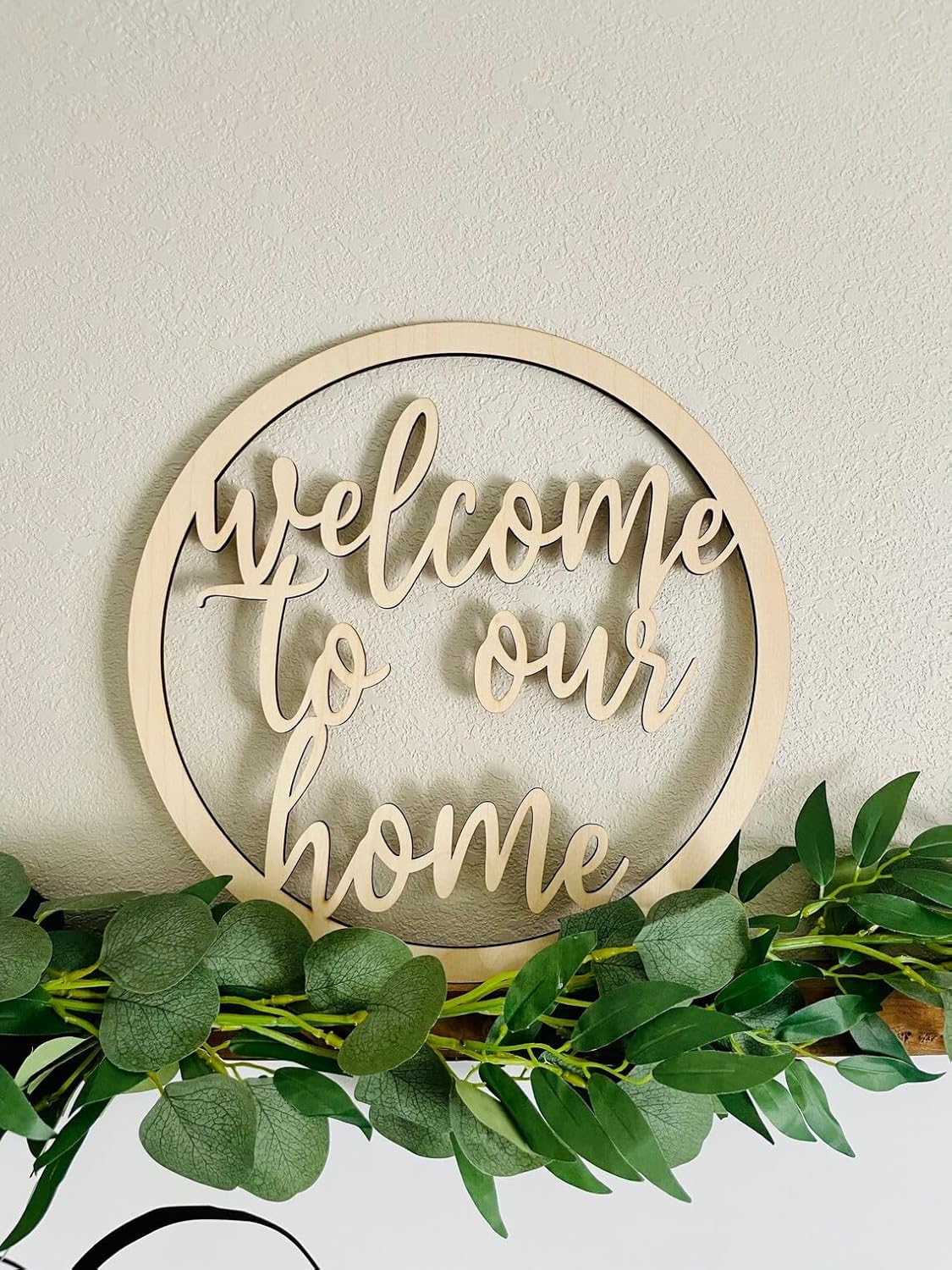 Wooden sign with 'welcome to our home' text surrounded by green foliage on a light background