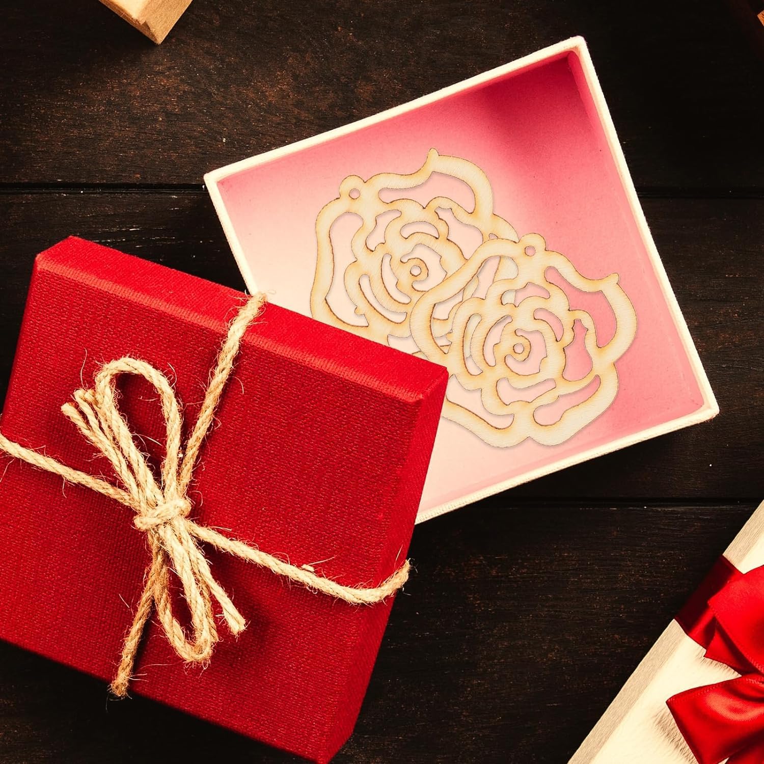 Red gift box with string bow next to a pink box with a gold rose design on a dark surface