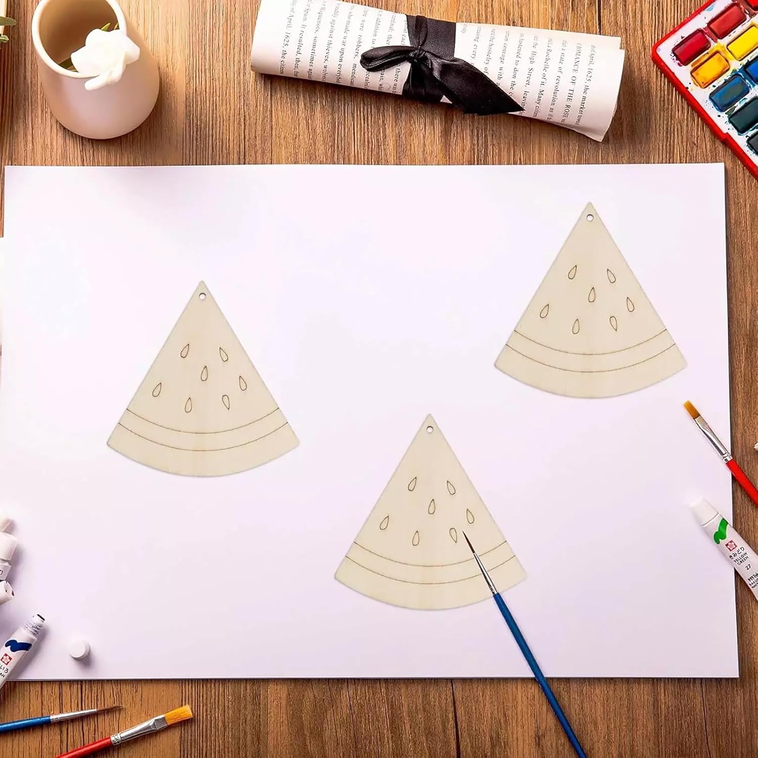 Three triangular watermelon slices on a piece of paper with painting supplies around.