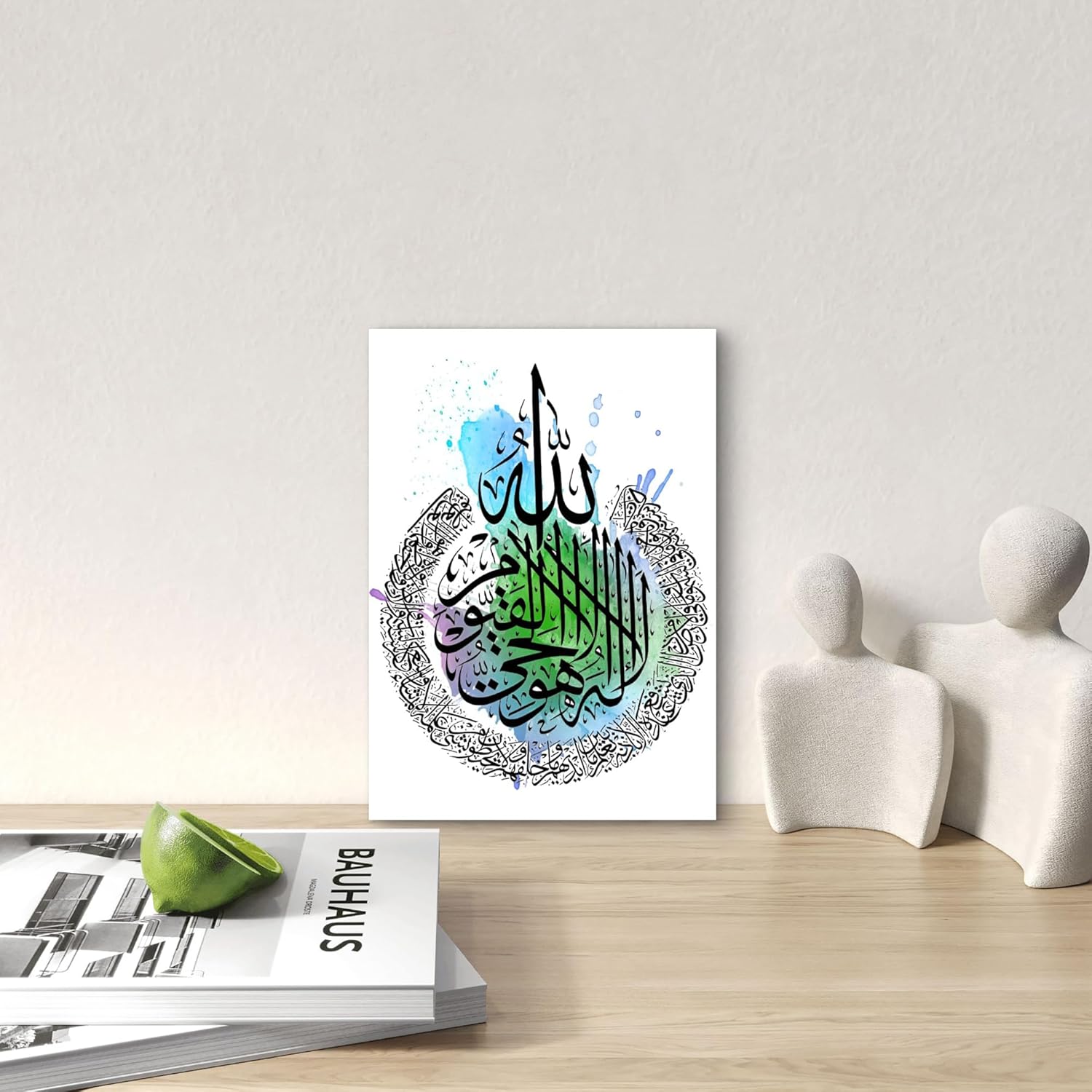 Decorative wall art with Arabic calligraphy on a white wall above a wooden surface with a green apple and magazine.