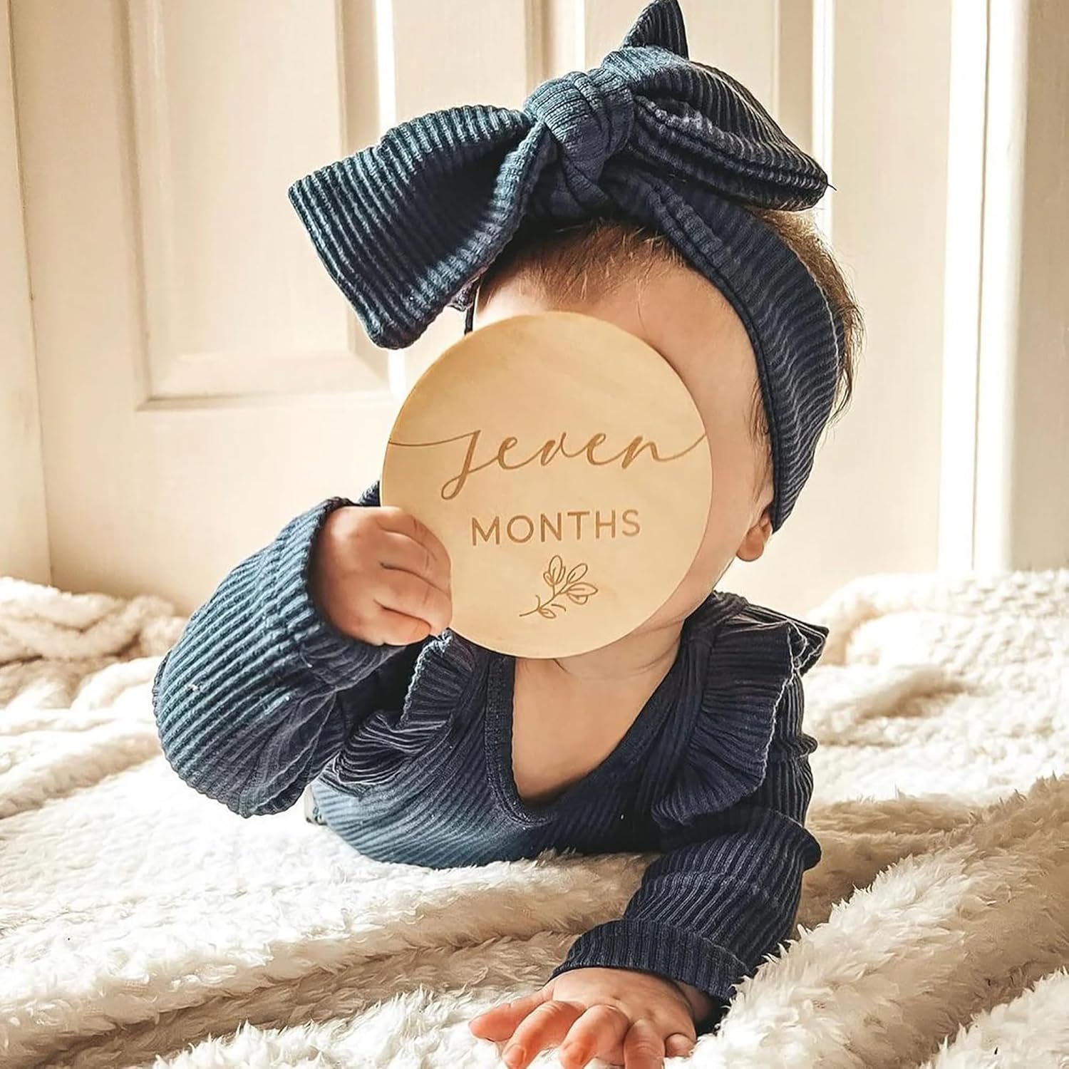 Baby wearing a blue headband and holding a 'Seven Months' sign on a soft surface.