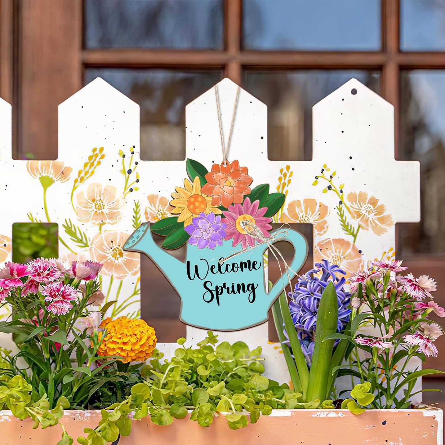 Decorative sign with flowers and a watering can saying 'Welcome Spring' in front of a white picket fence.