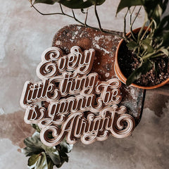 Wooden sign with 'Every little thing is gonna be alright' text on a textured surface with plants.
